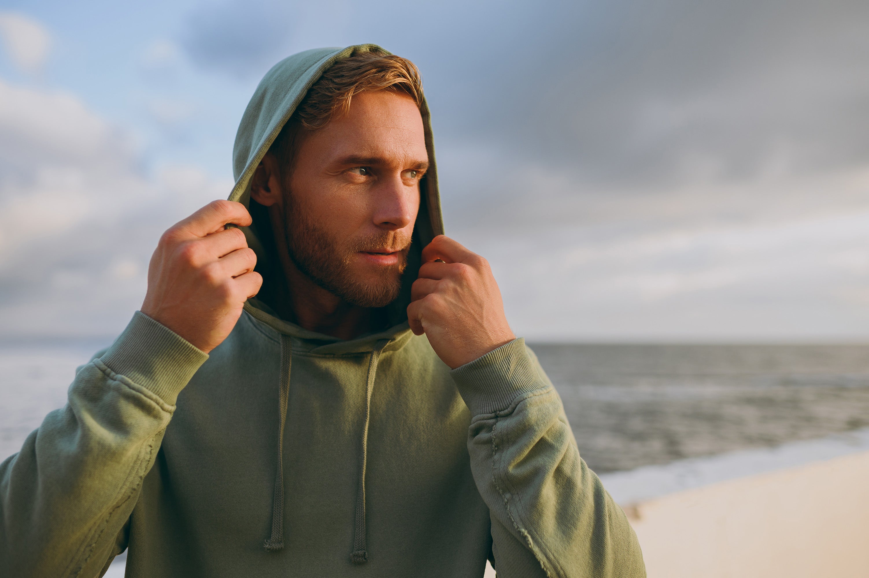 Man on the beach looking in the distance wearing a green hoodie pulled over the top of his head